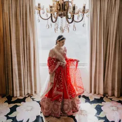 A classic bridal portrait in a beautifully lit room. The chandelier and elegant decor complement the bride's stunning red lehenga, creating a picture of pure grace.