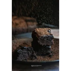A stack of rich, dark chocolate rum brownies being dusted with cocoa powder. The slow shutter speed captures the motion of the dusting, adding a dynamic and decadent feel to the image.