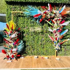 A vibrant and colorful take on boho decor. This setup uses a green hedge wall as a base, with a gold frame decorated with brightly dyed pampas grass and florals in shades of red, yellow, and blue for a festive, eclectic look.