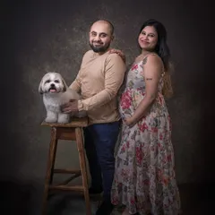 An expecting couple poses with their beloved Shih Tzu in a classic studio portrait. The dog sits proudly on a stool, an important member of the growing family.