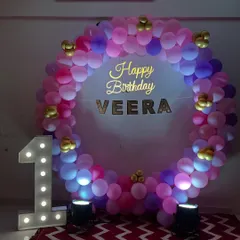 A circular balloon frame in shades of pink and purple for Veera's first birthday. The setup is completed with a "Happy Birthday" sign and a large light-up number '1'.