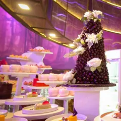 A breathtaking dessert landscape under dramatic purple light. Featuring a stunning grape tower, tiered cakes, and individually plated sweets, this is gourmet grandeur.