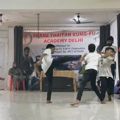 Action from an indoor training event. A student executes a powerful kick during a multi-person sparring drill, learning to manage threats from different directions.