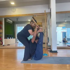 An instructor assists a student using a stick to achieve greater extension and core engagement in a challenging leg-lift variation.
