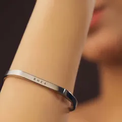 A close-up on a woman's arm, showing the delicate and clean engraving of the name 'RHEA' on a silver cuff. The minimalist design makes it perfect for daily wear.