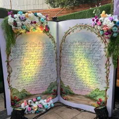 A unique and regal Bridgerton-themed Godh Bharai (baby shower) entrance, featuring a giant open storybook prop. The pages are filled with elegant calligraphy and the book is adorned with lush floral arrangements, creating a dramatic and romantic welcome.