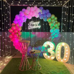 This vibrant 30th birthday celebration on a terrace features a rainbow-themed balloon arch and a 'Happy Birthday' neon sign. The setup is framed by a cube of fairy lights and includes large, illuminated '30' marquee numbers.