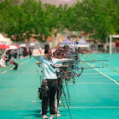 A photograph from the National Women's Archery Tournament in Ambaji, showing a line of archers taking aim. This image captures the scale of the event and the collective focus of the athletes.