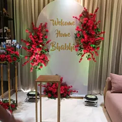 A stunning "Welcome Home Bhabhi" setup in vibrant red. The backdrop includes large floral bouquets and a personalized message in gold, creating a grand welcome.