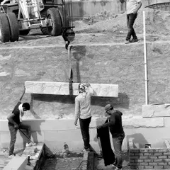 A black and white shot capturing the raw effort of construction on the House of Solid Stone. Workers guide a heavy stone lintel, lowered by a crane, into its final position, a moment of precision and teamwork.