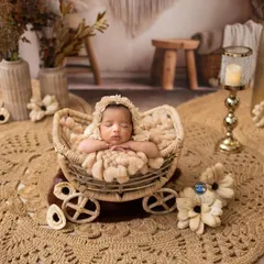 A newborn baby girl sleeps soundly in a wicker pram, wearing a delicate lace bonnet. The entire scene is styled with earthy, bohemian tones using jute rugs and dried flowers.