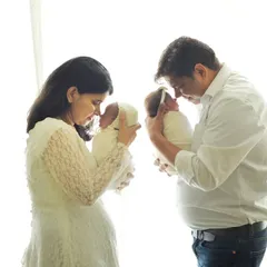 The first family portrait with twins is a milestone. Here, both parents hold one baby each, creating a balanced and emotional image against a soft, bright background.