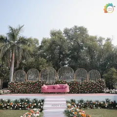 A full view of a beautiful outdoor stage setup. The backdrop features golden arches with hanging floral strings, a base of colorful flowers, and a chic pink velvet sofa, creating a fresh and modern look for a day event.