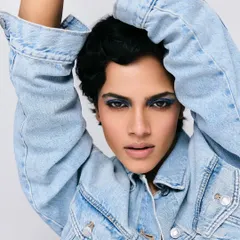 A striking portrait featuring a model with a pixie cut and bold blue smokey eyes. The styling is minimal, focusing on the denim jacket and the powerful, direct gaze.