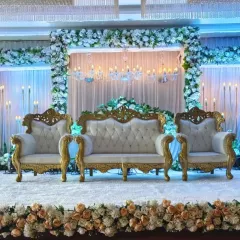 An opulent stage design at Ellora Banquet hall. This setup features a backdrop of white flowers and elegant chandeliers, paired with regal gold and white throne-style seating for a truly luxurious feel.