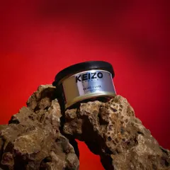 A bold shot of a men's grooming product, "Keizo Silver," placed on a rugged rock against a vibrant red background.