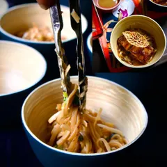 A collage showing the preparation of a noodle dish. Tongs lift a tangle of noodles into a bowl, ready to be combined with a rich sauce and toppings, showcasing the final steps of our live noodle bar.
