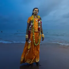 A devotee stands at the edge of the sea in Tiruchendur, the twilight sky creating a dramatic backdrop for his traditional attire and garlands.