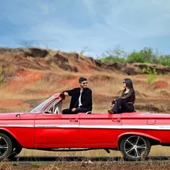 A wide-angle shot of the couple seated on a classic red convertible, set against a rugged, natural landscape for a dramatic pre-wedding portrait.