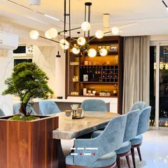 The dining area is designed for memorable meals, featuring a stunning marble-top table with a live bonsai centerpiece. The space is illuminated by a modern globe chandelier, with a custom bar cabinet in the background.