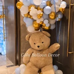 A close-up of a life-sized teddy bear holding a large bunch of balloons, a perfect welcoming prop for any celebration.