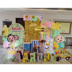 This Cocomelon first birthday setup features a vibrant backdrop with a gold shimmer panel, themed plinths, and colorful balloon arrangements. Large character cutouts and custom block letters for the name 'Aaruhi' complete the scene.