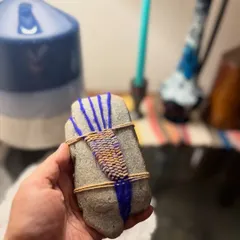 This is the finished woven pebble, held in hand to show its scale and texture. The combination of the hard, smooth stone and the soft, colorful yarn creates a unique tactile object that feels wonderful to hold.