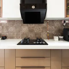 A close-up of a modern kitchen with a stylish patterned tile backsplash, a sleek black hob, and beige cabinets with elegant rose-gold handles.