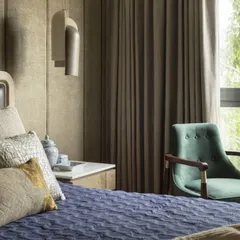 A serene bedroom at the Jor Bagh Residence with a view of the lush green outdoors. The design combines a textured upholstered headboard, a deep blue quilted bedspread, and a comfortable armchair to create a peaceful escape.