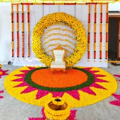 A vibrant and traditional puberty function setup, featuring a massive floral rangoli on the floor made from marigold petals. The backdrop includes a full floral ring and a classic white chair, creating a perfect photo opportunity.