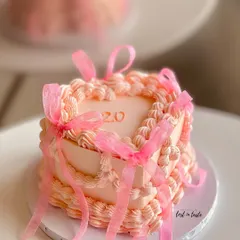 A beautiful heart-shaped cake in a soft pink hue, decorated with classic vintage Lambeth piping and delicate organza ribbons. This design is perfect for celebrating a 20th birthday in style.