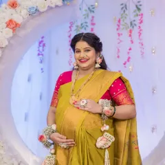 A beautiful solo maternity portrait of the mother-to-be, cradling her baby bump. She stands before a floral moon-gate, her traditional yellow saree and floral accessories perfectly complementing the festive baby shower decor.