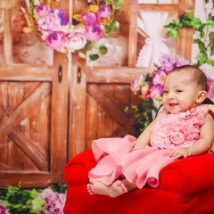 This little one is all smiles, sitting comfortably in a miniature red armchair. The floral backdrop adds a touch of rustic charm to her sitter portrait.