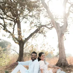 A happy couple sits back-to-back on a picnic blanket under trees, smiling brightly. The warm sunlight filtering through the leaves creates a beautiful, relaxed atmosphere for this outdoor shoot.