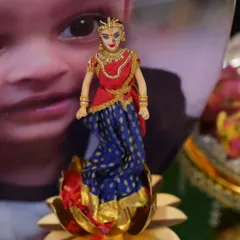 A close-up of a doll dressed for a puberty ceremony, placed in a brass lotus holder. The backdrop features a photo of the girl being celebrated.