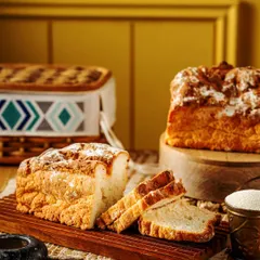 A rustic loaf of artisan bread, sliced to show its soft interior. The warm, home-style setup with a wooden board and picnic basket evokes a feeling of comfort and traditional baking.
