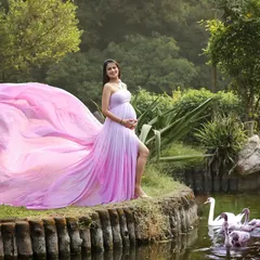 A scene from a fairytale. The mom-to-be looks like a princess in her flowing pink gown, standing by a pond with swans in a beautiful, lush park.