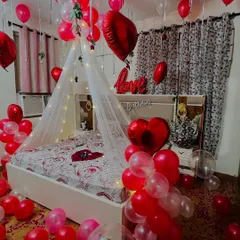 I design romantic room surprises for anniversaries and special moments. This setup features a fairy light canopy over the bed, with red and white balloons, heart-shaped foil balloons, and rose petals to create a truly memorable experience.