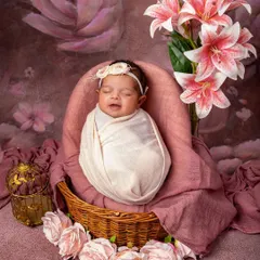 Elegance in full bloom. A smiling newborn is swaddled and settled in a basket, surrounded by soft pink roses and lilies. The floral backdrop adds a touch of classic, painterly beauty.