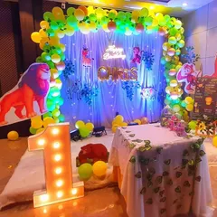 A vibrant jungle-themed first birthday setup. The decor includes a backdrop with a leafy curtain, animal cutouts like a lion and deer, and a light-up number '1'.