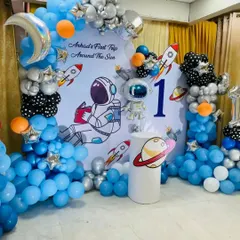 A "First Trip Around The Sun" space-themed birthday party. This creative setup features a custom backdrop with an astronaut, balloon galaxies in blue, black, and silver, and props like rockets and planets.