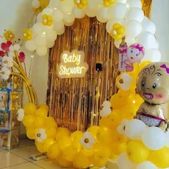 A bright and cheerful baby shower decoration in yellow and white. This circular backdrop combines a shimmering gold curtain with a custom balloon arrangement, a neon sign, and cute baby-themed foil balloons.