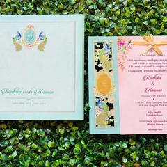 A beautiful pastel wedding invitation featuring a traditional peacock and Ganesha motif. The card has a laser-cut panel with gold foil accents and a ribbon tie, blending classic Indian design elements with a soft, modern color scheme.
