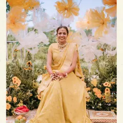 The bride, radiant in yellow, seated for her Haldi ceremony. The backdrop of giant, ethereal white and yellow paper flowers and a field of sunflowers creates a stunning and artistic portrait moment.