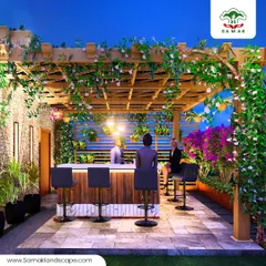 An evening view of a terrace we designed as a social hub. The space features a custom wooden pergola draped in flowering vines, a fully functional bar, and varied lighting to create a warm, inviting atmosphere for entertaining guests.