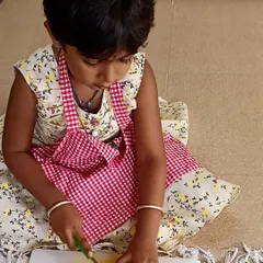With great concentration, a child practices slicing mangoes. These hands-on kitchen activities build confidence and introduce basic food preparation skills in a safe, guided way.