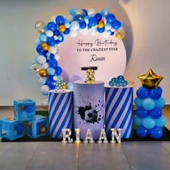 A sporty football-themed birthday party in blue, white, and gold. The decor features a circular backdrop, striped plinths with a football graphic, and star-shaped balloon accents.