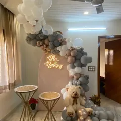 This is an example of a stylish baby shower decoration designed for a home setting. The setup includes a modern arched backdrop, an "Oh Baby" neon sign, and a sophisticated balloon arrangement in grey, taupe, and white, complete with a teddy bear.