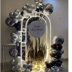 A very glamorous and modern party setup. This features a lit archway filled with pampas grass, surrounded by black, silver, and chrome balloons and disco ball accents for a nightclub vibe.
