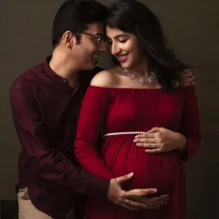 An expecting couple shares a loving gaze in this intimate fine art portrait. The deep red of her dress contrasts beautifully with the muted green backdrop, highlighting their connection.